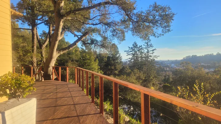 Finished wood-and-cable railing along an unstained deck beside a house, overlooking trees and a distant valley.