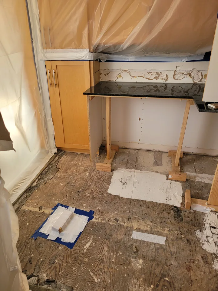 Kitchen during repair with protective plastic, exposed flooring, opened cabinetry, and a temporarily supported countertop.